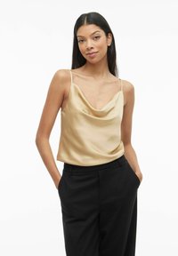 Soeva kaelusega satiinist kampsun sooja beežiga, õhukeste rihmadega, koos mustade kohandatud pükstega, näidates sujuvat tekstuuri ja elegantset langevust.