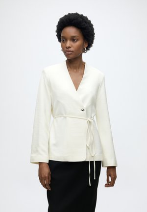 Kobieta w białej kopertowej kurtce z wiązaniem z boku i czarnej spódnicy, stojąca na jednolitym jasnym tle.