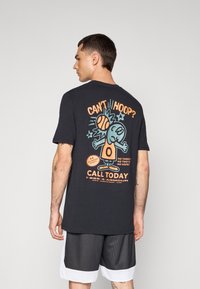 Sort bomulds T-shirt med et farverigt cartoon grafik på ryggen, med teksten "KAN DU IKKE SPILE?" og et basketball motiv. Korte ærmer.