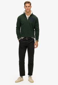 Zielony polarowy sweter z kołnierzem na pół-zamek, szara koszula pod spodem, czarne spodnie i beżowe sneakersy. Posiada haftowane logo na piersi.