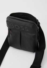 Sac en nylon noir à bandoulière avec une finition texturée. Comprend une fermeture éclair sur le dessus, deux plis avant et un logo. Inclus une bandoulière tissée robuste.