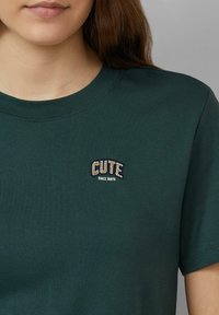 Donkergroene T-shirt met een ronde halslijn, voorzien van een gestikte patch waarop "CUTE" in lichte en donkere kleuren staat, met daaronder "SINCE BIRTH".