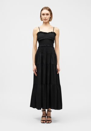 Γυναίκα που φοράει μακρύ μαύρο φόρεμα χωρίς μανίκια, με στρώσεις και με σφηκωμένη κορώνα και λεπτές τιράντες, σε συνδυασμό με μαύρα σανδάλια με τακούνι και φαρδιά λουράκια στο αστράγαλο.