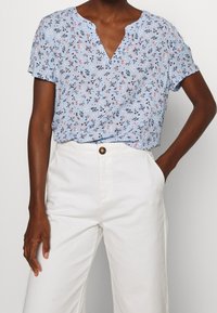 Blaue kurzärmlige Bluse mit V-Ausschnitt. Verziert mit kleinen rosa und schwarzen Mustern. Kombiniert mit weißen, hoch-taillierten Hosen.