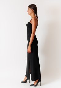 Abito nero senza maniche con corpino aderente, dettagli arricciati e uno spacco laterale, abbinato a scarpe nere con tacco a punta.