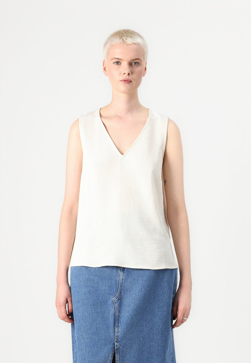 Top sans manches en V crème, avec un tissu doux et texturé et une coupe décontractée. Associé à une jupe en denim bleu clair avec une fente centrale.