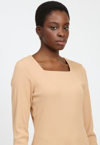 Camiseta de manga larga color beige con escote cuadrado, textura acanalada y diseño ajustado. Tejido suave con costuras mínimas y una silueta clásica.