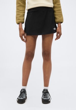 Naine, kes kannab musta North Face lühikest seelikut, beežikat fliisjaki, valgeid sokke ja musti matkajalatsid, seisab valge tausta ees.