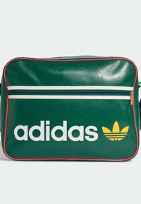 Zielona winylowa torba z białym suwakiem, czerwonym obszyciem i dużym białym logo „adidas”. Na froncie widoczny charakterystyczny żółty emblemat koniczyny. Gładka tekstura.