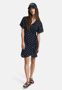 Marineblauwe jurk met polkadots, met korte mouwen en een gerimpelde zoom. Gecombineerd met bruine sandaaltjes met banden en een marineblauw pet met witte tekst.