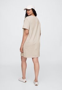 Béžové bavlnené šaty v štýle trička s krátkymi rukávmi, voľným strihom a hladkou textúrou. Doplnkom sú biela čiapka a svetlé tenisky.