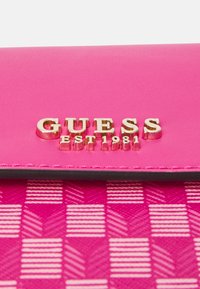 Roza novčanica od kože s teksturiranim uzorkom šahovnice, s ušivenim zlatnim logotipom "GUESS EST 1981" i kontrastnim crnim šavovima.