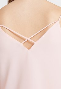 Světle růžový top s designem zkřížených ramínek na zádech, měkkou texturou a V-výstřihem.