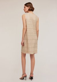 Beiges ärmelloses Kleid mit einem strukturierten, geschichteten Design, verziert mit Paillettenakzenten und Fransen am Saum. Glattes Material mit lockerer Passform.