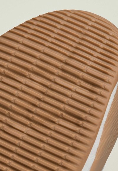 Rubber shoe sole in tan, featuring a textured pattern of raised stripes and grooves for grip; edge is white with a smooth finish.