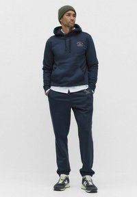 Mężczyzna w granatowej bluzie z kapturem i pasujących dresach, w białych trampkach i zielonej wełnianej czapce, stojący z rękami w kieszeniach.