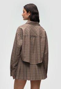 Brązowa koszula w kratę w oversize'owym stylu z wyraźnym kołnierzykiem, opadającymi ramionami i zaokrąglonym dołem, wykonana z miękkiego materiału o teksturowanym wykończeniu.