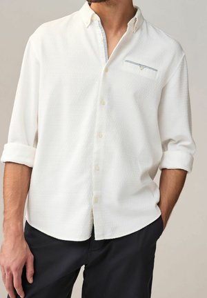 Homme portant une chemise à manches longues blanche texturée avec les poignets retroussés et un pantalon noir, main dans la poche.