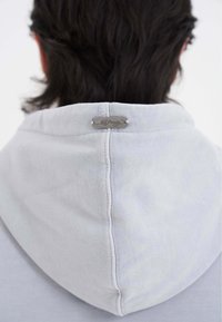Felpa grigia chiara con un'etichetta metallica sul collo. Il tessuto è morbido e ha una texture liscia, e il design include un cappuccio.