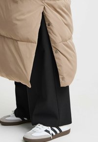Cappotto puffo beige con bottoni a pressione, abbinato a pantaloni neri. Sneakers bianche con dettagli grigi e strisce nere. Texture liscia e design imbottito.