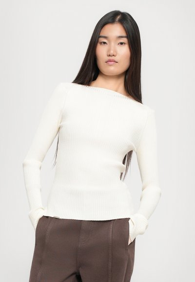 Ribbed, off-shoulder top in cream with long sleeves. Soft texture, fitted design, and a straight hem. Paired with brown pants.