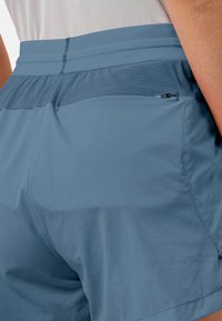 Close-up van blauwe sportshorts met een textuur in de tailleband en een klein met rits sluitend zakje op de rechter heup, gedragen met een witte top.