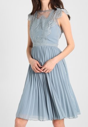 Mujer con un vestido azul claro sin mangas, con detalles de encaje en el corpiño y una falda plisada, manos juntas en la cintura.