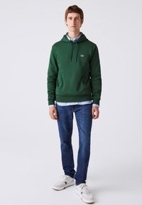 Joven de pie, vistiendo una sudadera verde, jeans azules, zapatillas blancas y una camisa de cuadros azul por debajo, sobre un fondo blanco liso.