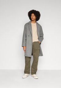Grijze wollen jas met een dubbelzijdig ontwerp, gecombineerd met een beige t-shirt en olijfgroene cargobroeken. Witte sneakers maken de outfit af.