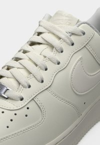 Sneaker in pelle bianca con superficie strutturata, punta rotonda e prominente swoosh laterale. Presenta lacci piatti e dettagli con occhielli in metallo.