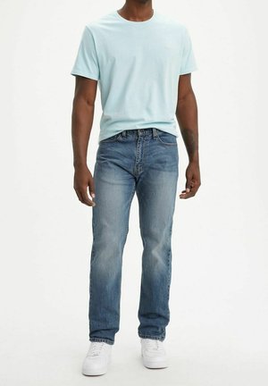 Man wearing light blue short-sleeve t-shirt, blue denim jeans, and white sneakers, standing against a plain white background.