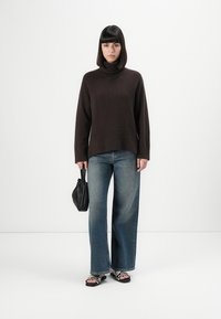 Femme se tenant devant un fond blanc, portant un pull col roulé marron foncé, un jean évasé, des sandales à motif et tenant un sac à main noir.