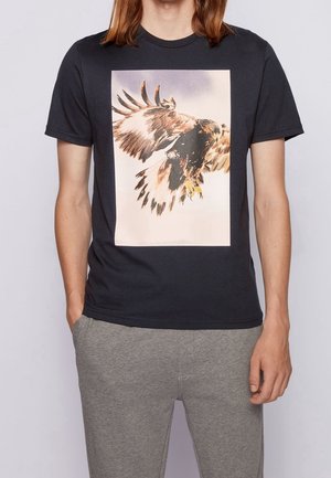 Person, die ein schwarzes T-Shirt mit einem großen Grafik eines fliegenden Adlers und hellgraue Hose trägt, steht mit den Händen in den Taschen.