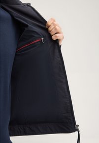 Osoba trzymająca otwartą czarną kurtkę, pokazując wewnętrzną kieszeń z czerwonym zamkiem błyskawicznym na jednolitym jasnym tle.