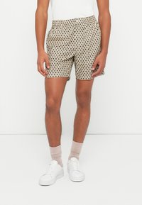 Uomo che indossa pantaloni corti beige a fantasia, scarpe da ginnastica bianche, calzini beige chiaro e una maglietta bianca, in piedi contro uno sfondo bianco semplice.