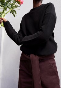 Pull noir côtelé à manches longues, associé à un pantalon marron avec un détail de taille nouée. Le mannequin tient un feuillage vert et des fleurs roses.