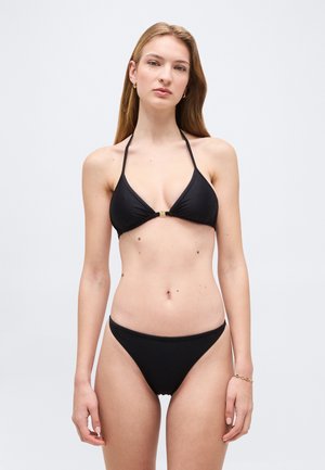 Femeie cu păr lung castaniu deschis, purtând un bikini negru în două piese, în picioare pe un fundal simplu gri deschis.