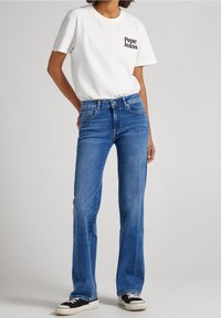 Vit bomullströja med svart "Pepe Jeans"-logotyp, kombinerad med blå utsvängda denimjeans och svarta sneakers med vita sulor.