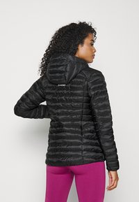 Doudoune noire avec capuche, présentant des sections matelassées horizontales et un logo dans le dos. Portée avec des leggings roses.