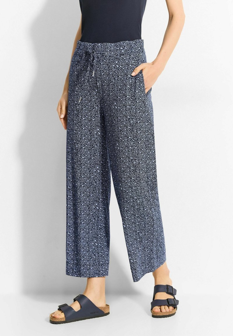 Patterned wide-leg trousers in navy with white floral designs. Lightweight fabric, drawstring waist, and side pockets. Paired with dark sandals.