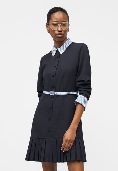 Ženska oblečena v črno plisirano obleko z modro-belim črtastim ovratnikom, manšetami in ujemajočim tankim pasom, stoji pred enobarvnim ozadjem.