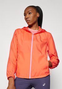 Veste coupe-vent légère orange avec une capuche, fermeture éclair sur le devant et poignets élastiques. Présente un logo discret sur la poitrine.