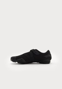 Zapatilla deportiva negra hecha de material flexible, con cierre de tiras, suela texturizada con protuberancias para agarre y un diseño aerodinámico.