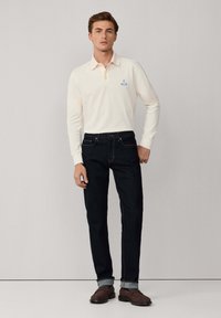 Polo a maniche lunghe color crema chiara con colletto, logo blu, abbinato a jeans in denim scuro. Modello che indossa scarpe in suede marroni.