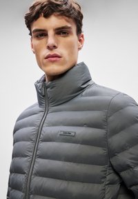 Tumšs pelēks, piepildīts, ar rāvējslēdzēju, jaciņa ar augstu apkakli. Iezīmēts ar horizontālām šuvēm un nelielu melnu etiķeti ar "Calvin Klein" uz krūtīm.