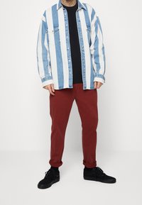 Camisa vaquera de rayas azul claro y blanco sobre una camiseta negra, combinada con pantalones rojo oscuro y zapatillas negras, posando sobre un fondo blanco.