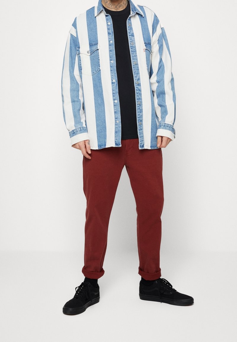 Camisa vaquera de rayas azul claro y blanco sobre una camiseta negra, combinada con pantalones rojo oscuro y zapatillas negras, posando sobre un fondo blanco.