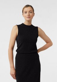 Kobieta z blond włosami w bezrękawnikowej czarnej połyskującej bluzce i spódnicy, stojąca z jedną ręką na biodrze na jednolitym białym tle.