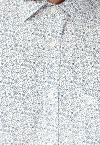 Biała koszula z małym niebieskim wzorem w kwiaty, z zapięciem na guziki i klasycznym kołnierzykiem. Materiał wydaje się lekki i fakturowany.