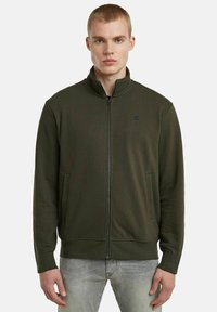 Felpa verde oliva con zip e colletto alto, tasche laterali, polsini a coste e un discreto logo sul petto. Tessuto misto cotone.
